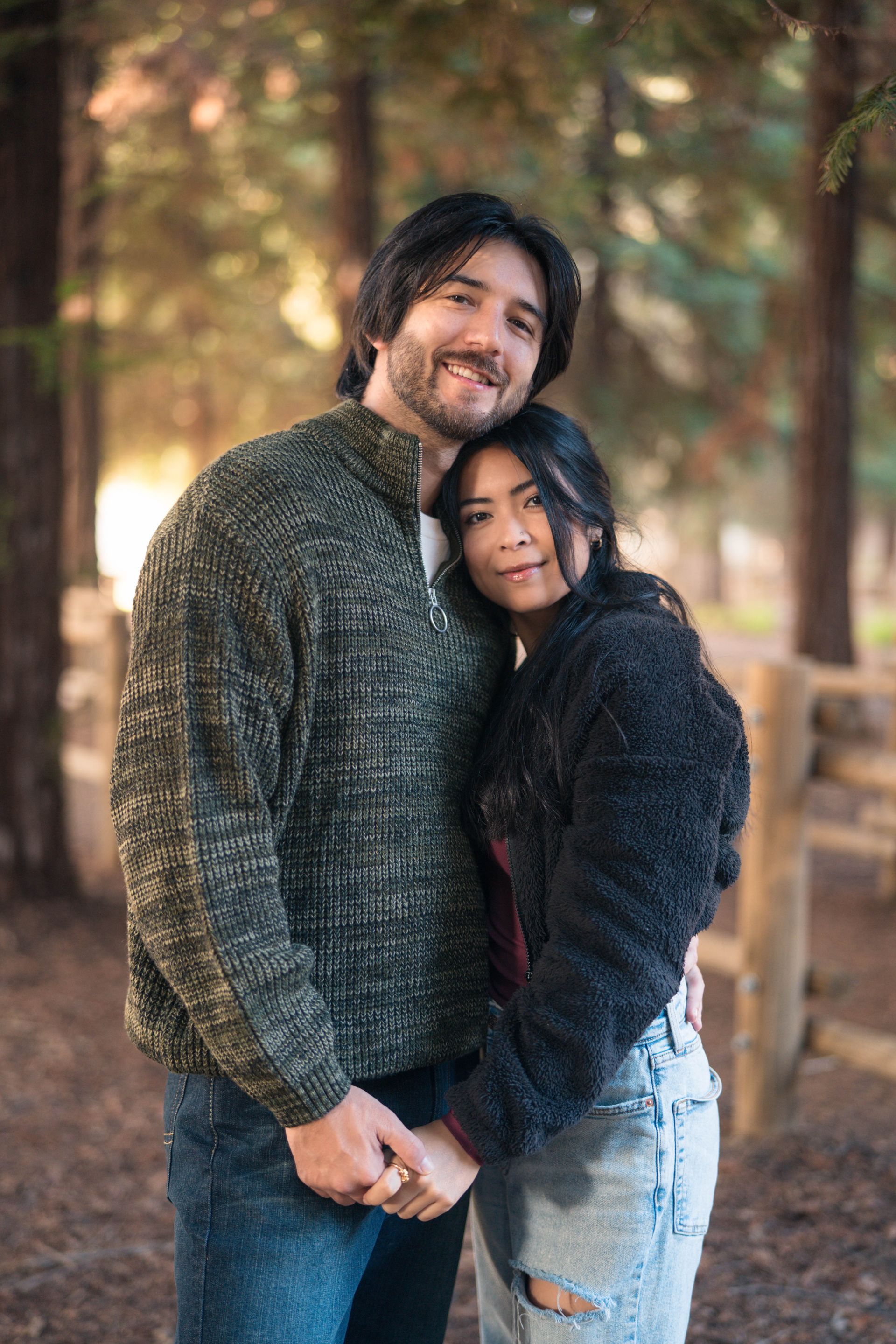 Couple embraces outdoors, holding hands. Man in green sweater, woman in black jacket; woods in background by Jelena's Photo