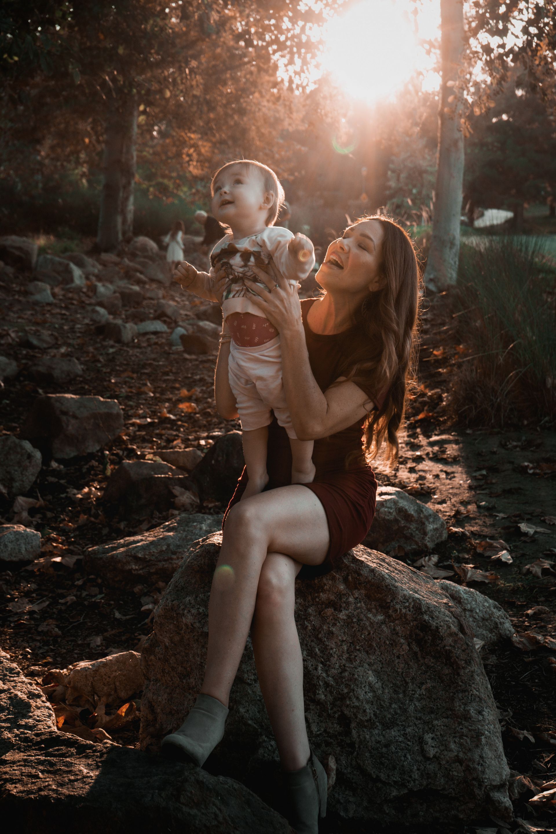 Woman holding baby in park, both smiling, bathed in sunlight by Jelena's Photo.