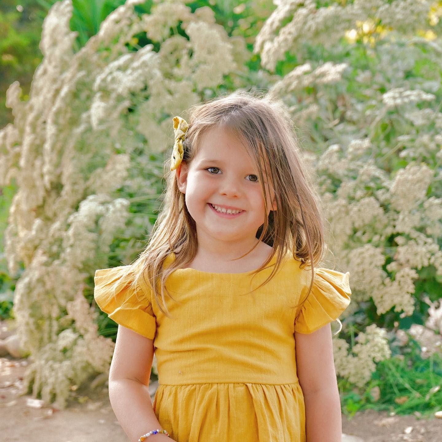 Girl in yellow dress smiles, standing in front of white flowers Jelena's Photo.