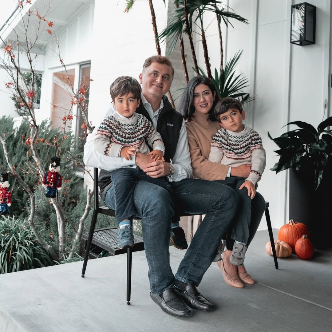 Family of four sitting on a chair outside, two children on parents' laps, pumpkins, and holiday decor.