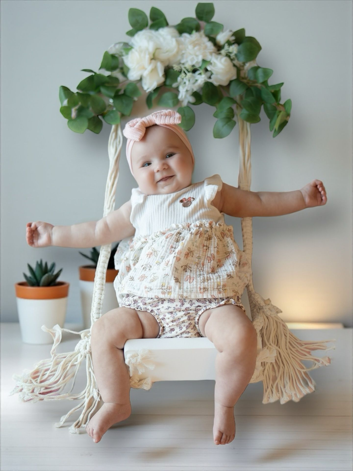 Baby in floral outfit on swing, smiling with arms outstretched; decorated with flowers and greenery Jelena's Photo.