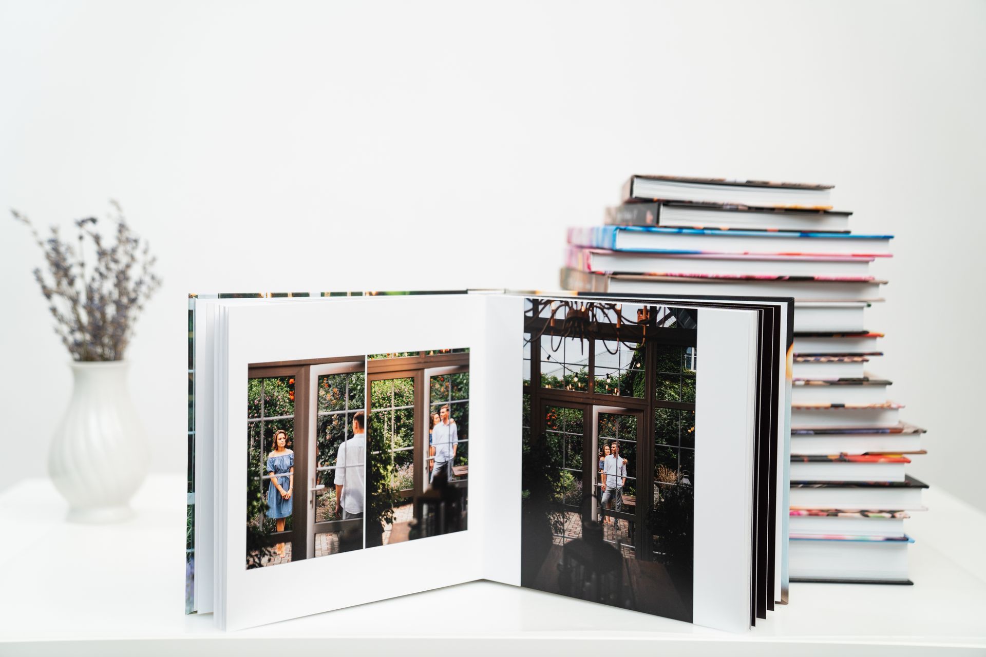 Open photo album with images; stack of albums in background; vase of flowers on table.