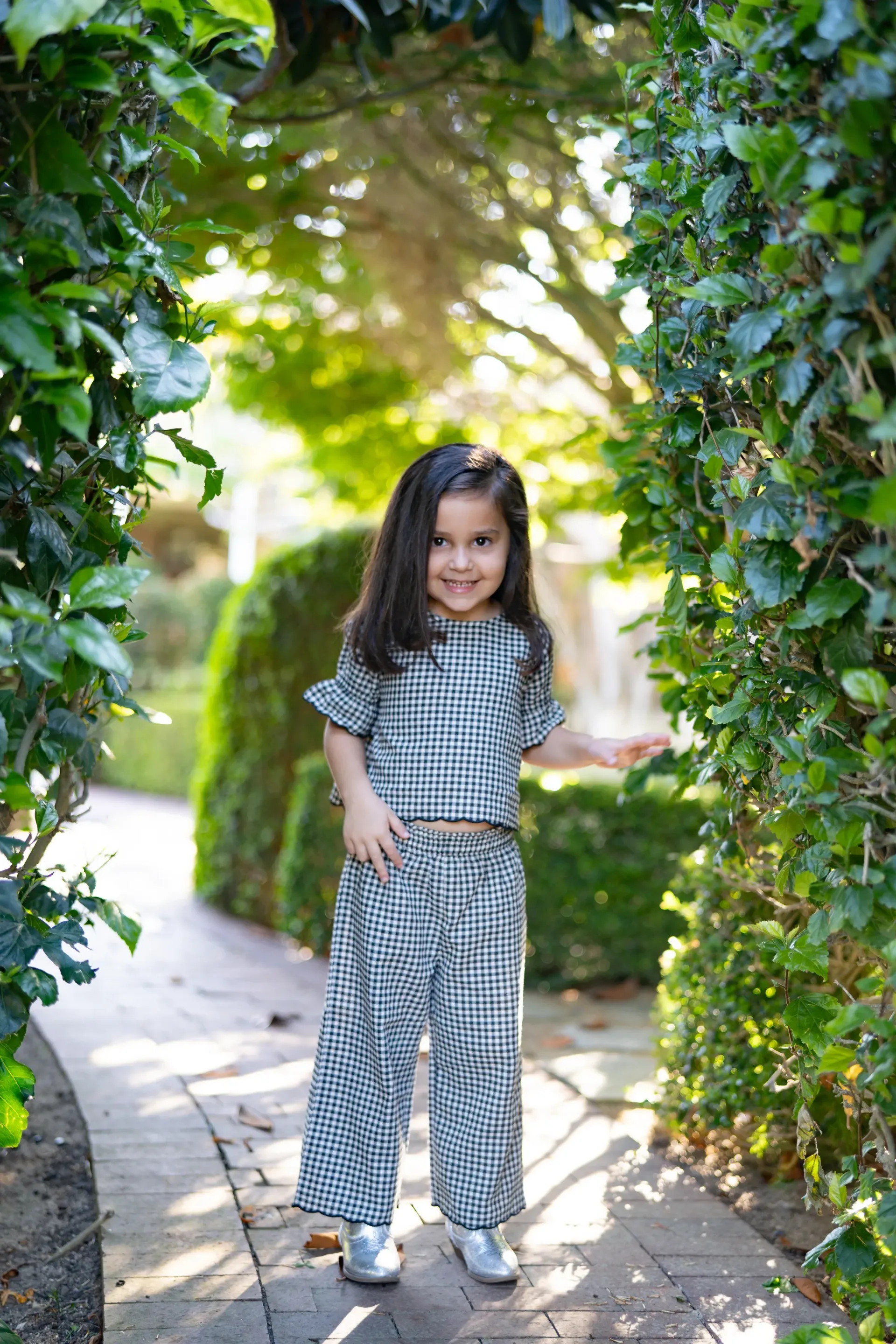 Young girl in checkered outfit stands smiling in a garden archway Jelena's Photo.