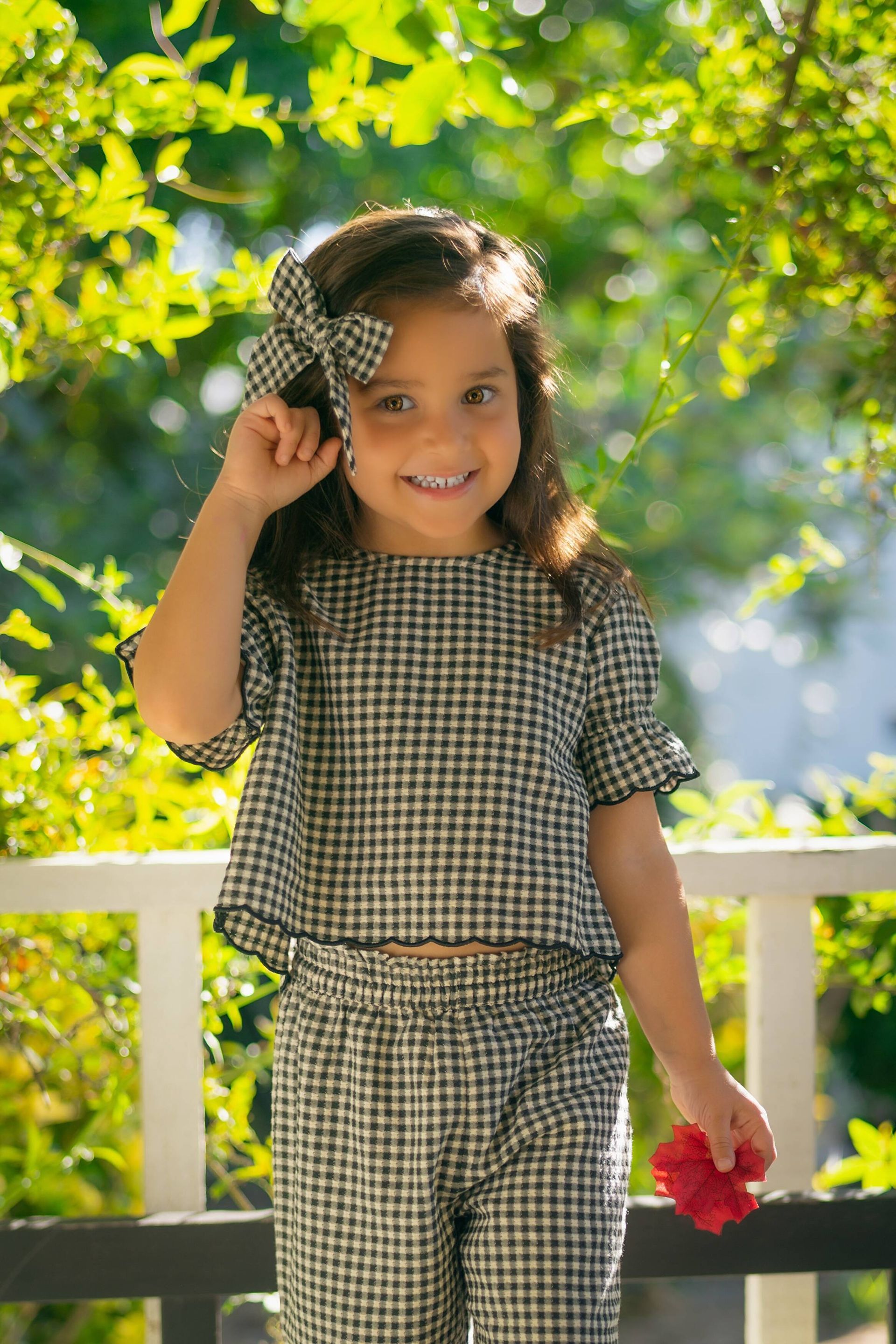 Girl in gingham outfit smiles outdoors, with bow in hair, holding red flower by Jelena's Photo.