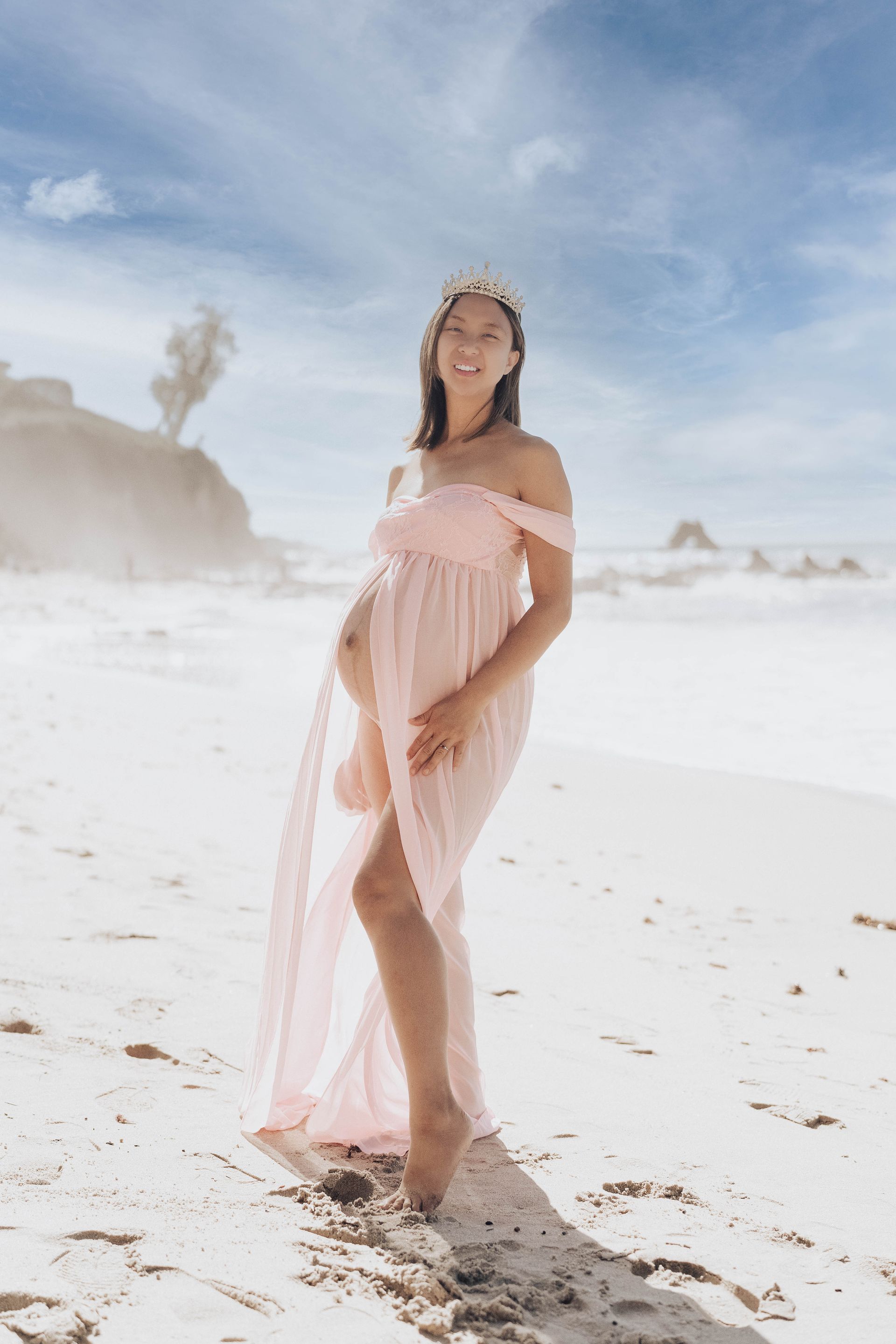 Pregnant woman in a pink dress on a beach, hand on her belly. Maternity photography by Jelena's Photo.