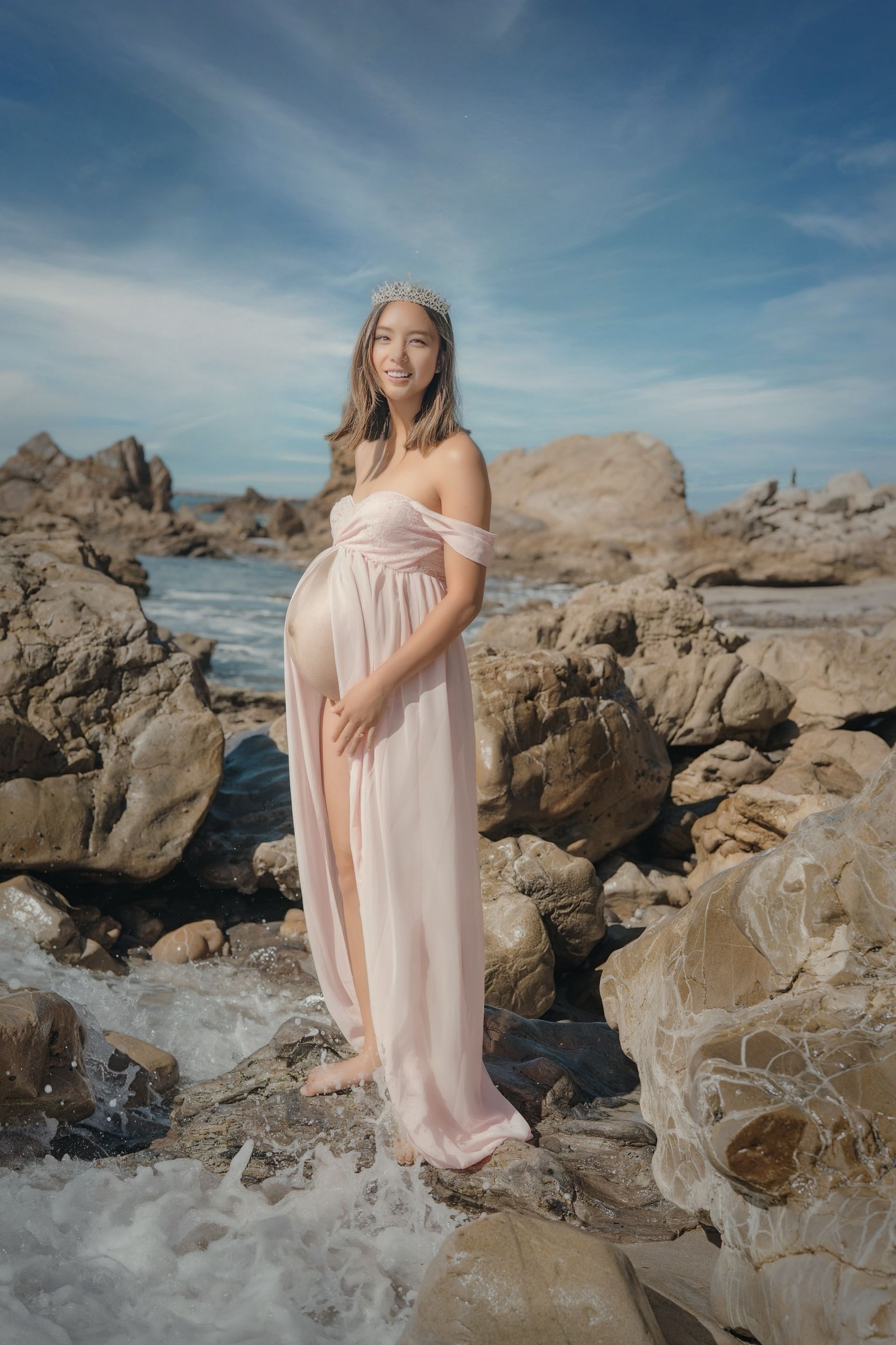 Pregnant person wearing a pink gown stands on rocks near the ocean, smiling. Maternity photography by Jelena's Photo.
