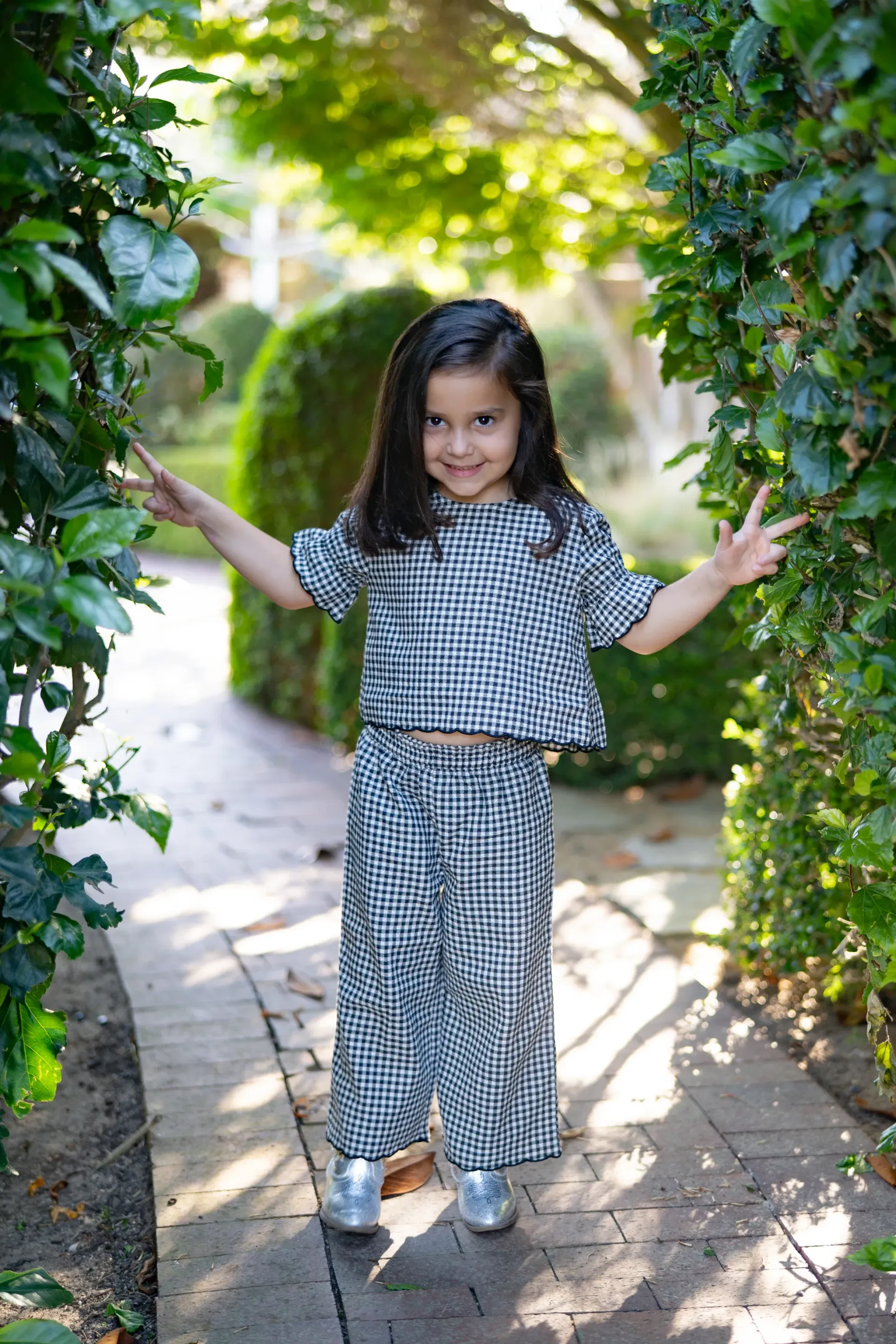 Young girl in gingham outfit smiles, arms open, standing on path, surrounded by greenery by Jelena's Photo.