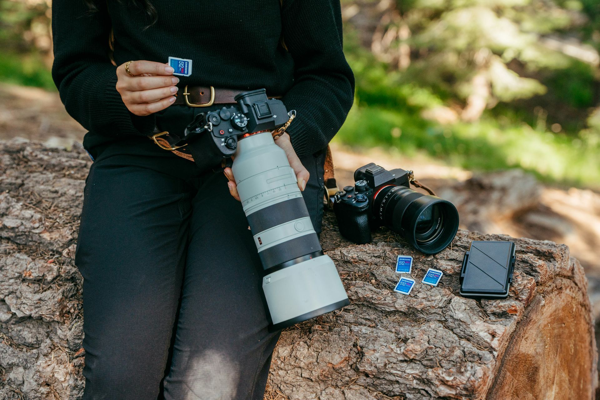 Photographer holding a memory card, with two cameras and a lens on a log.
