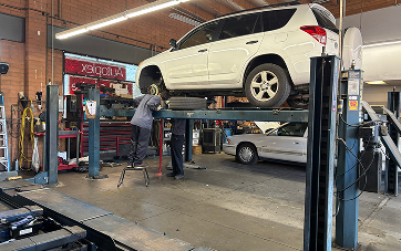 White SUV on a car lift in a repair shop as a mechanic works on a tire. | Autoplex Auto Service