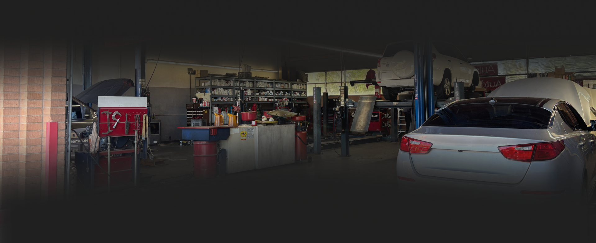 Inside a garage, a white car being worked on, other cars lifted on hoists, tools and shelves visible. | Autoplex Auto Service