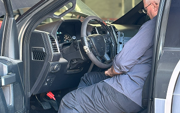 Person in a vehicle, sitting in driver's seat.  Car interior is dark.  The person wears a dark blue shirt and glasses.