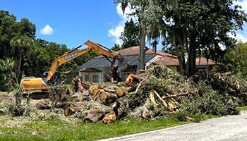 an excavator is moving a pile of logs in front of a house .