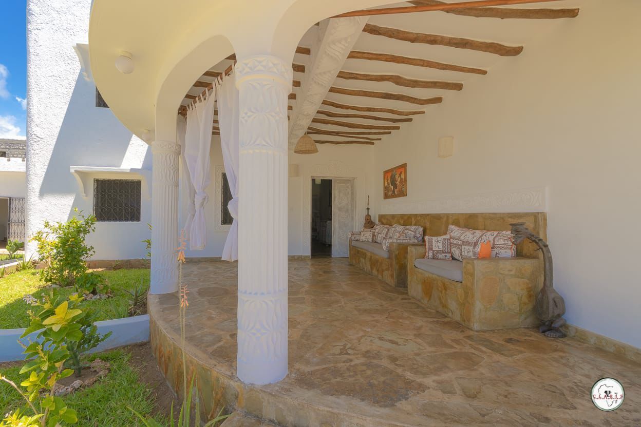 Covered stone patio with white columns, built-in bench seating, and curved wooden beams beside a house