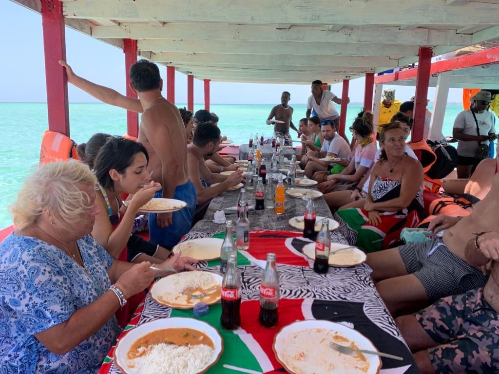 People dining together at a seaside table under a shaded canopy, with drinks and plates on a red tablecloth