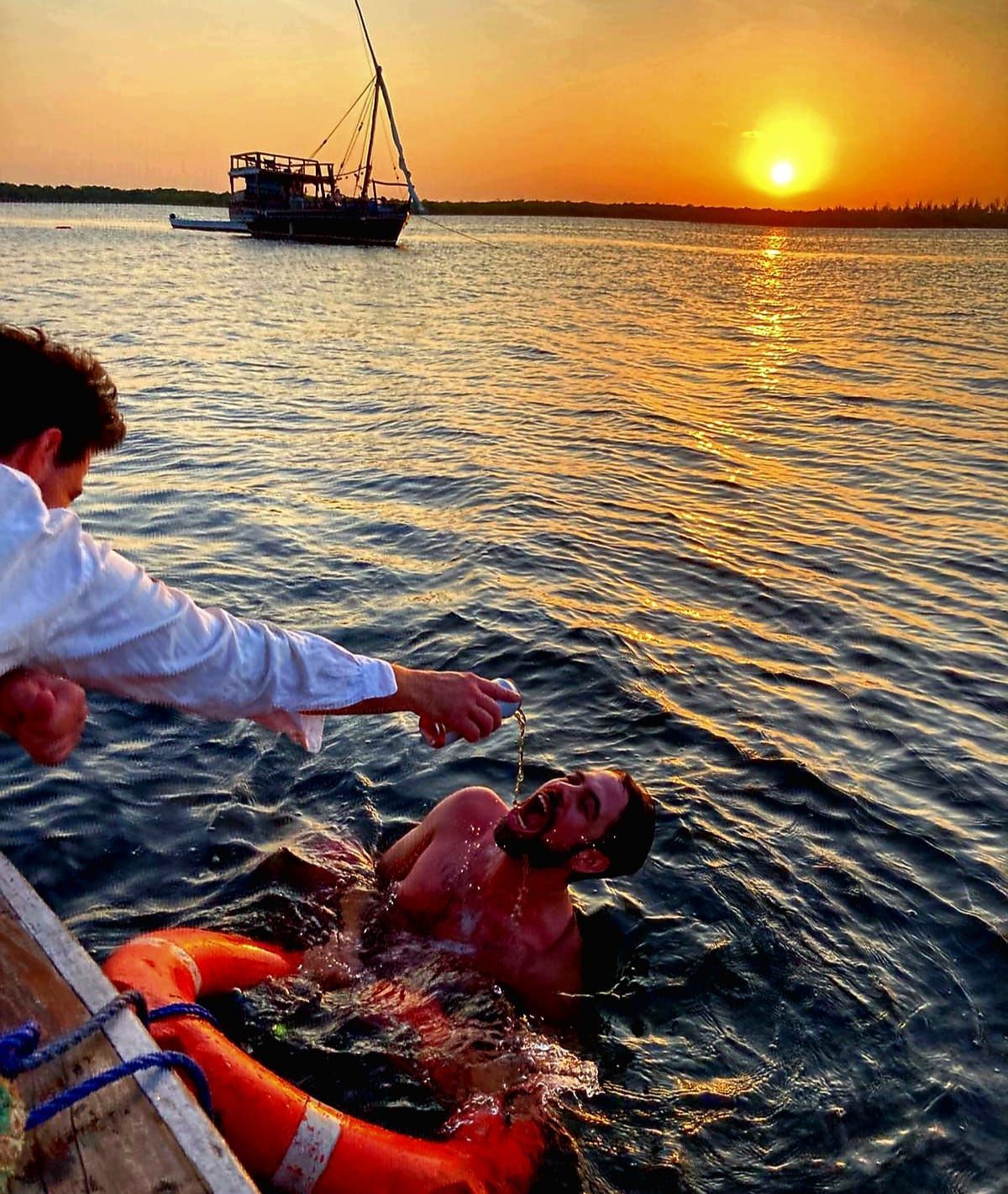 Person in life jacket in water at sunset, reaching toward someone on a boat; fishing boat in the distance.