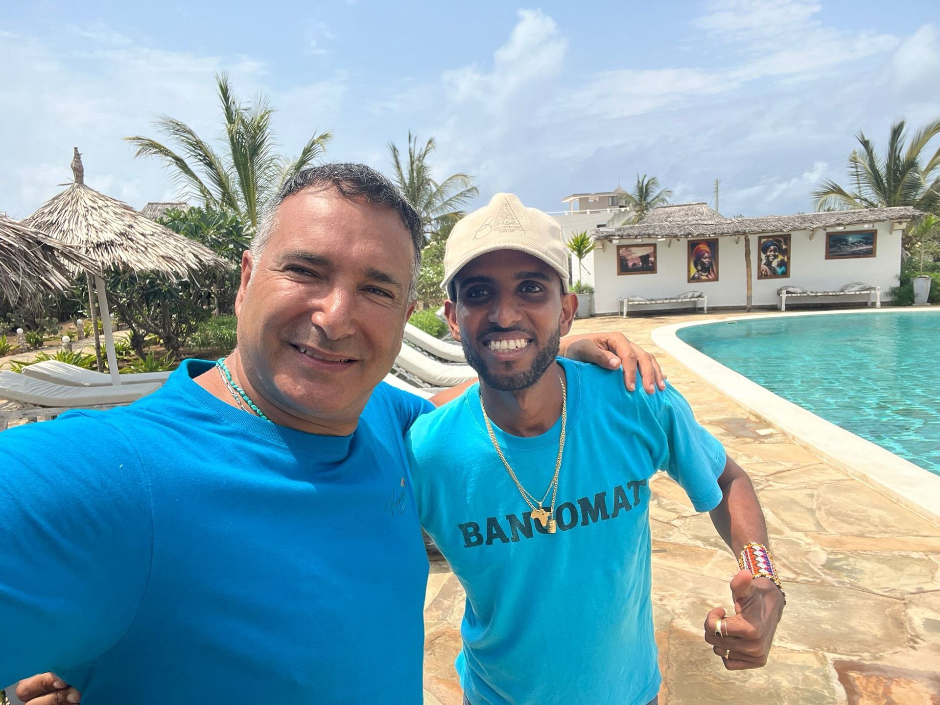 Two smiling people posing by a pool at a sunny resort, one in a blue shirt and one in a tan hat.