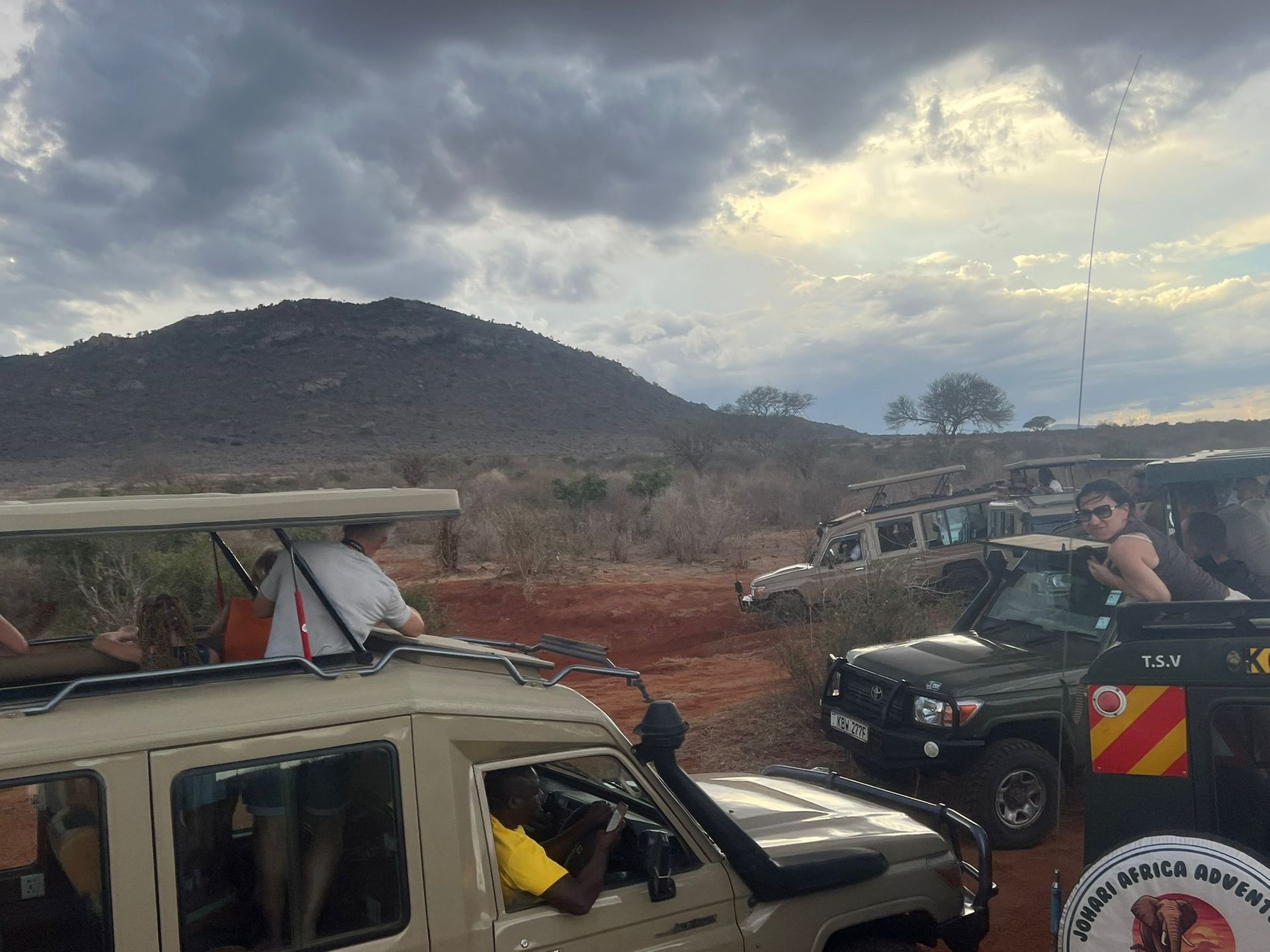 Vehicles on a muddy road near a hillside, with people gathered under a cloudy sky.
