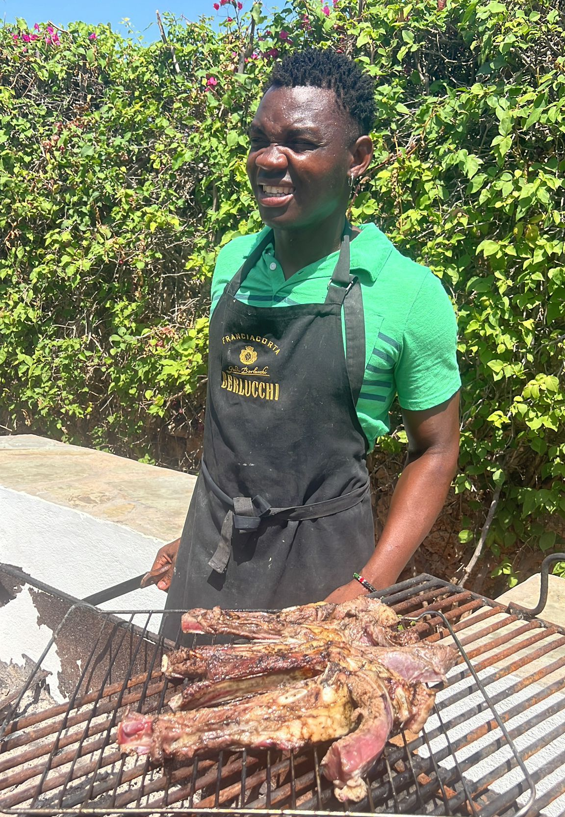 Man grilling large crab outdoors beside a green hedge, wearing a black apron and green shirt.