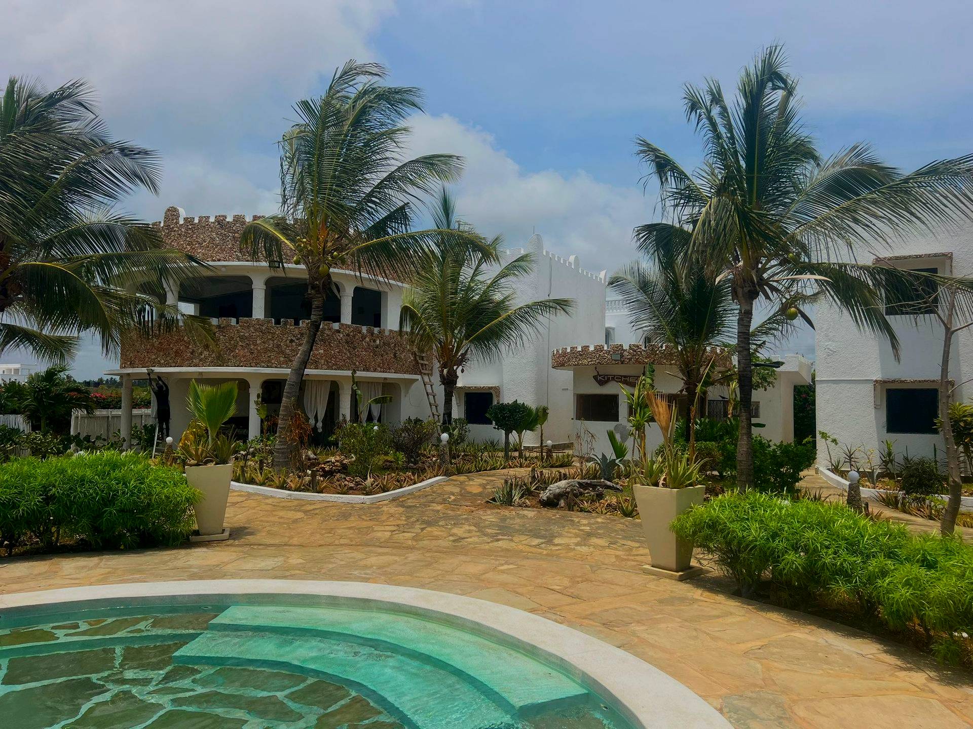 Tropical resort courtyard with a turquoise pool, palm trees, and white buildings under a blue sky