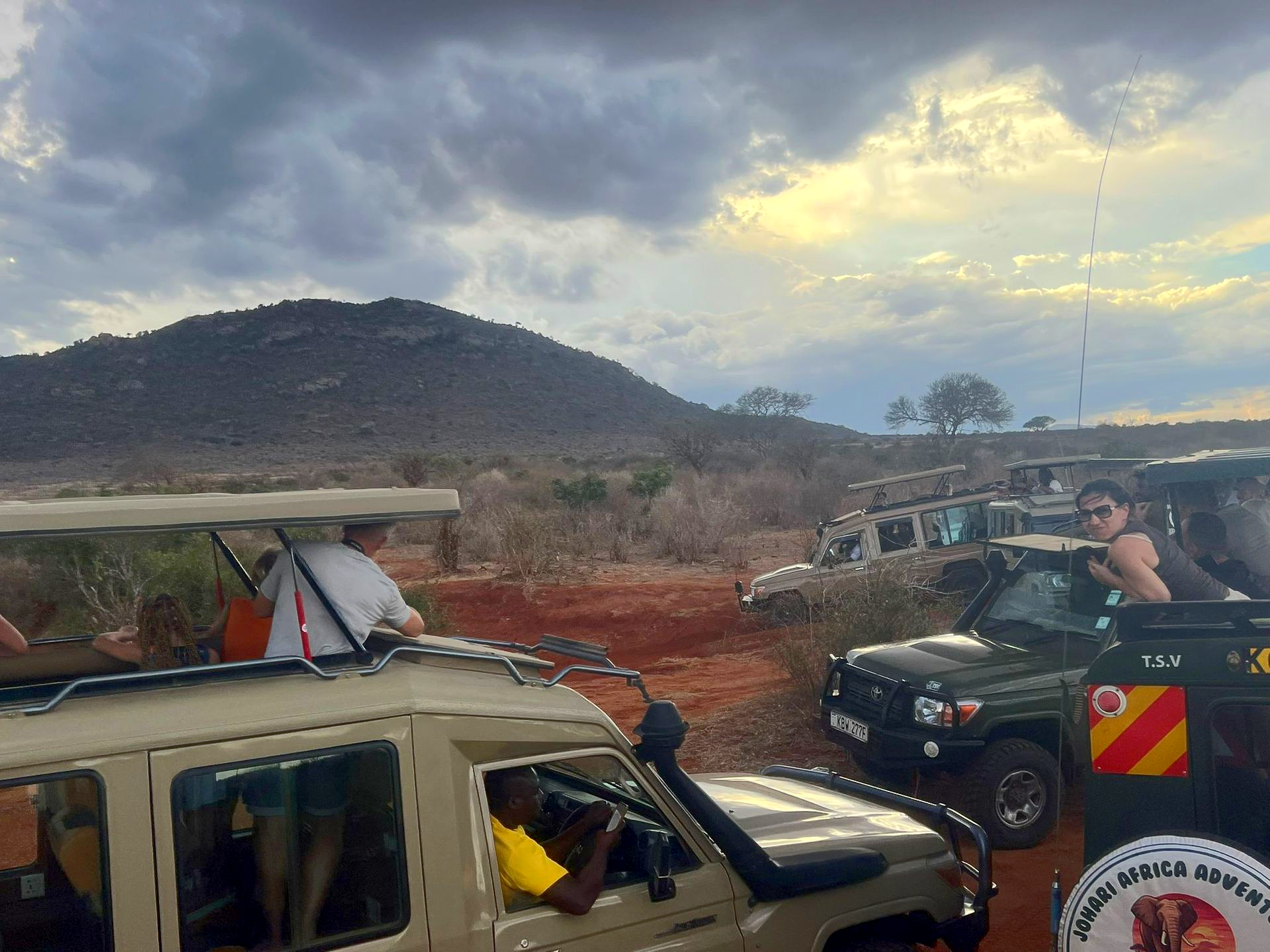 Safari vehicles on a dirt road near a hill under cloudy skies