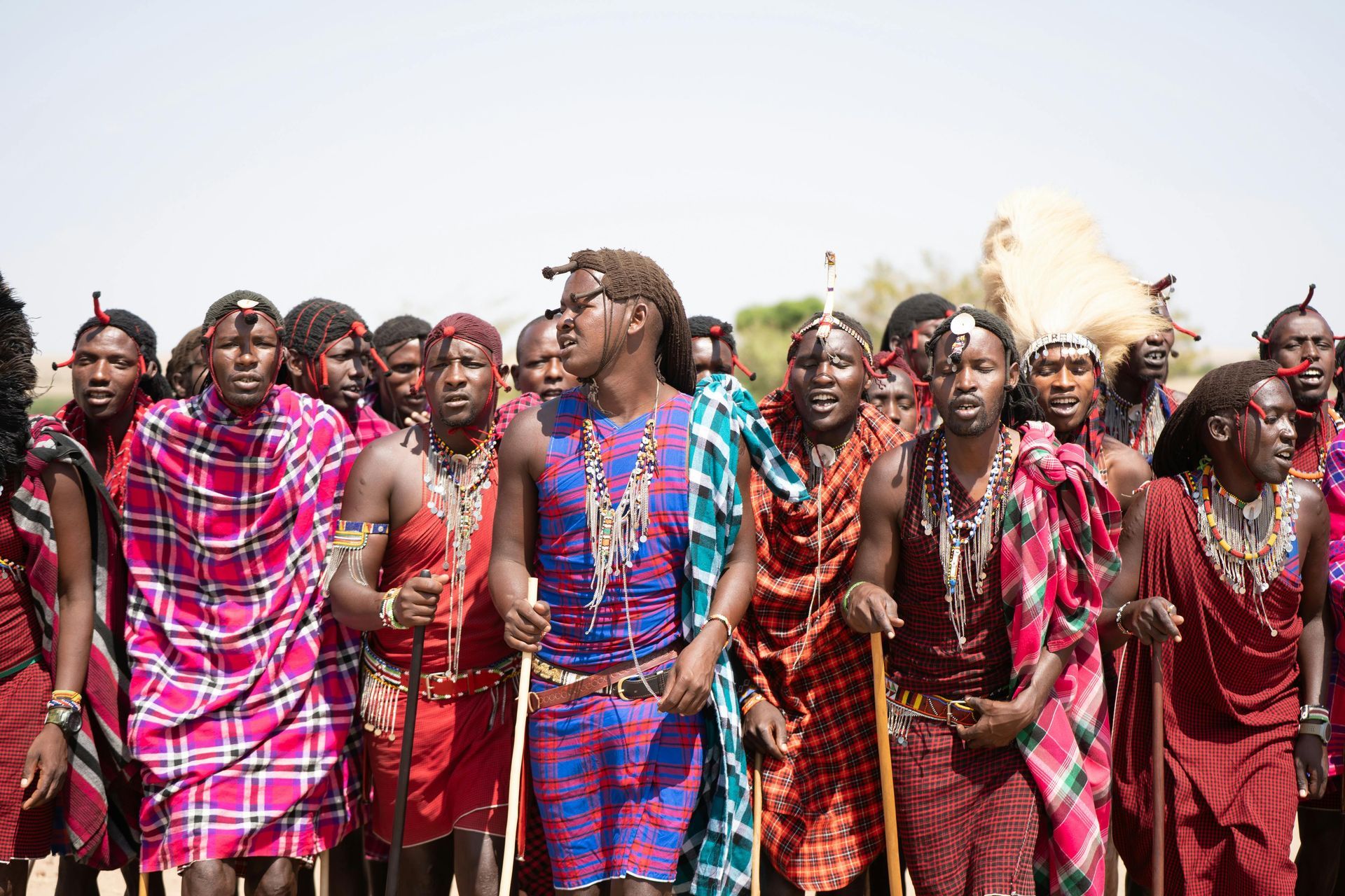 Group of people in colorful traditional attire standing together outdoors, some holding spears and feathered decorations