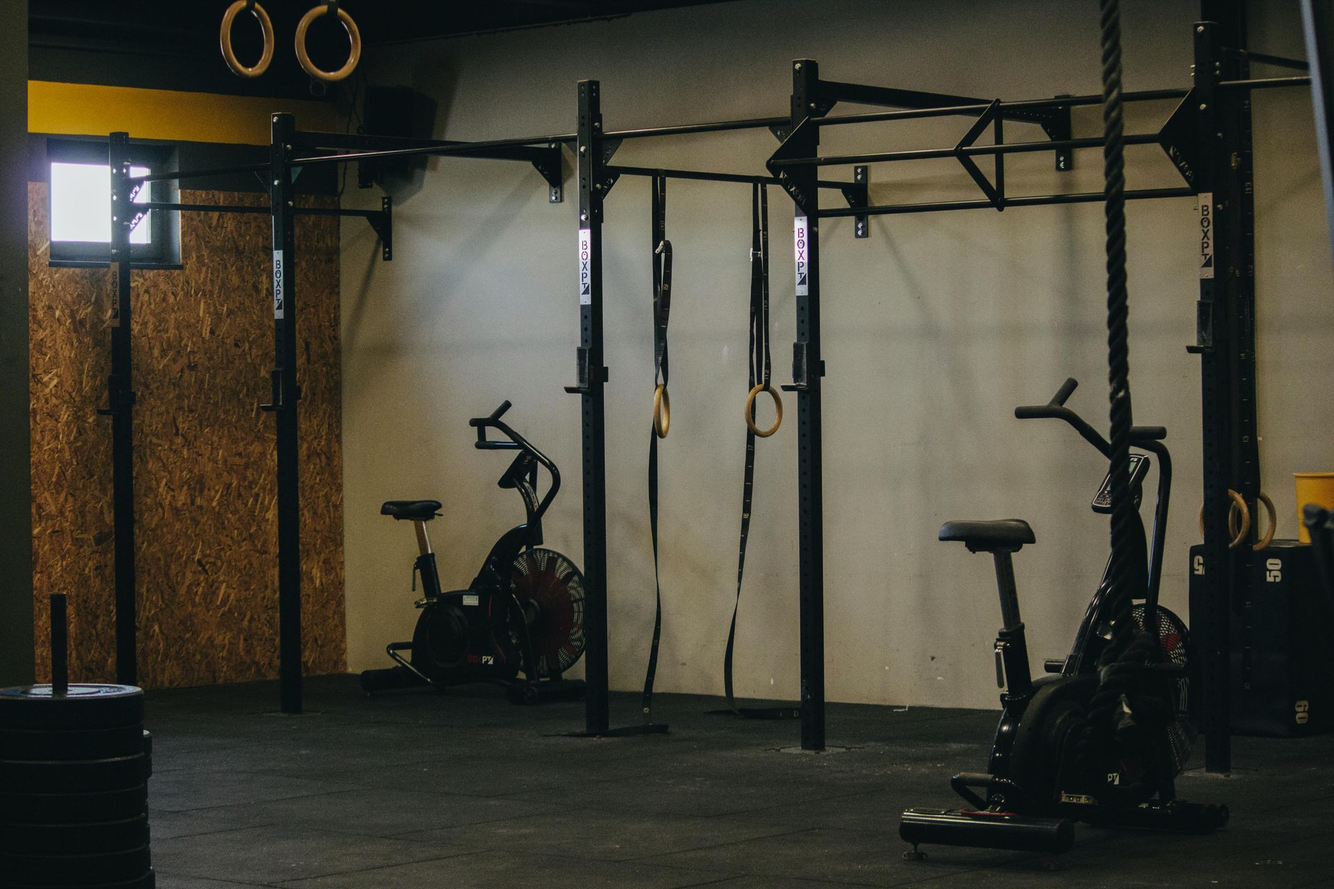 Dim gym with pull-up rig, exercise bikes, and wall-mounted rings on a dark floor.