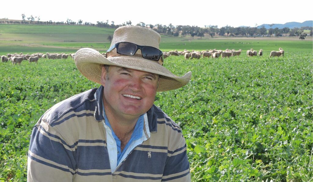 Smiling farmer in a straw hat standing in a green crop field with livestock grazing in the background