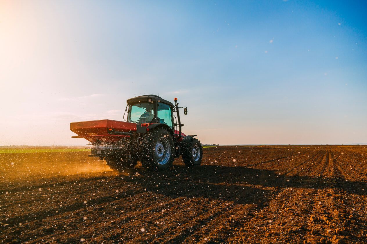 Tractor driving across a plowed field at sunset, spraying dust under a clear blue sky.