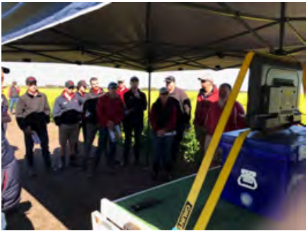 Group of people standing under a canopy beside a blue machine with a yellow beam outdoors