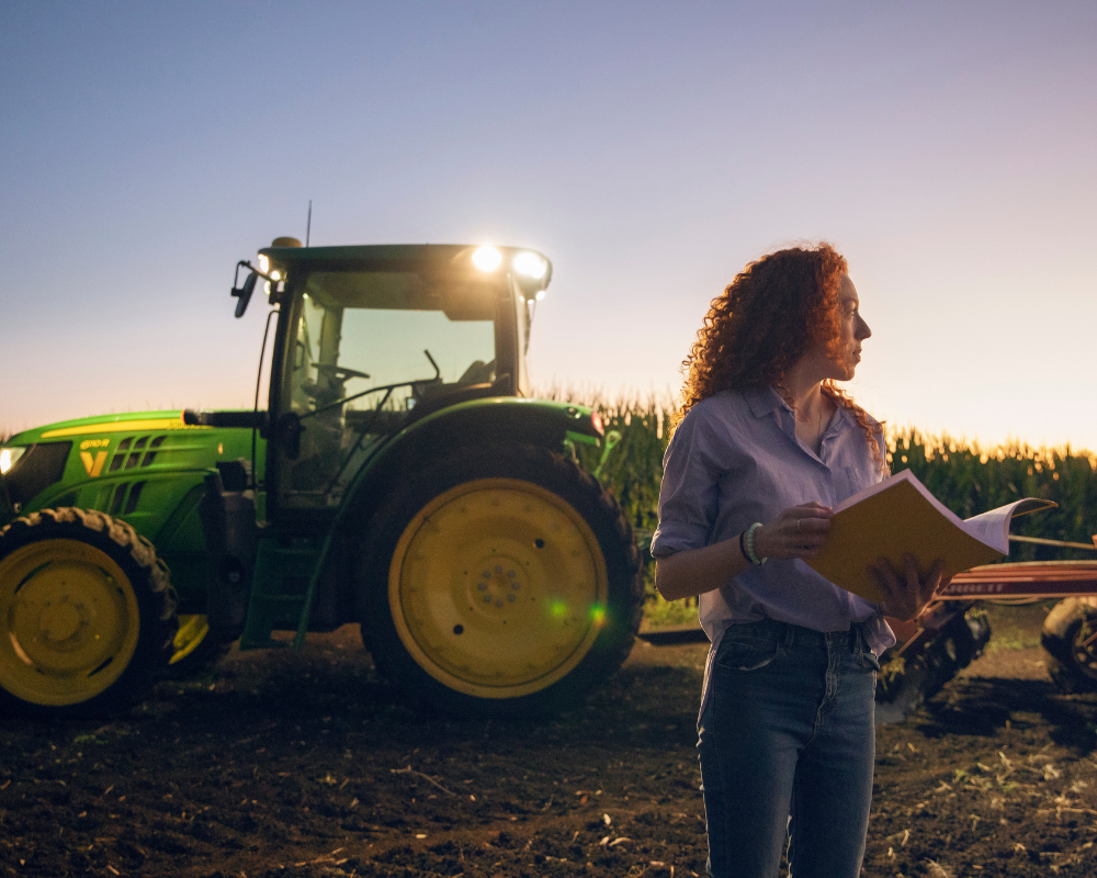 Woman holding a tablet beside a green tractor in a field at sunset