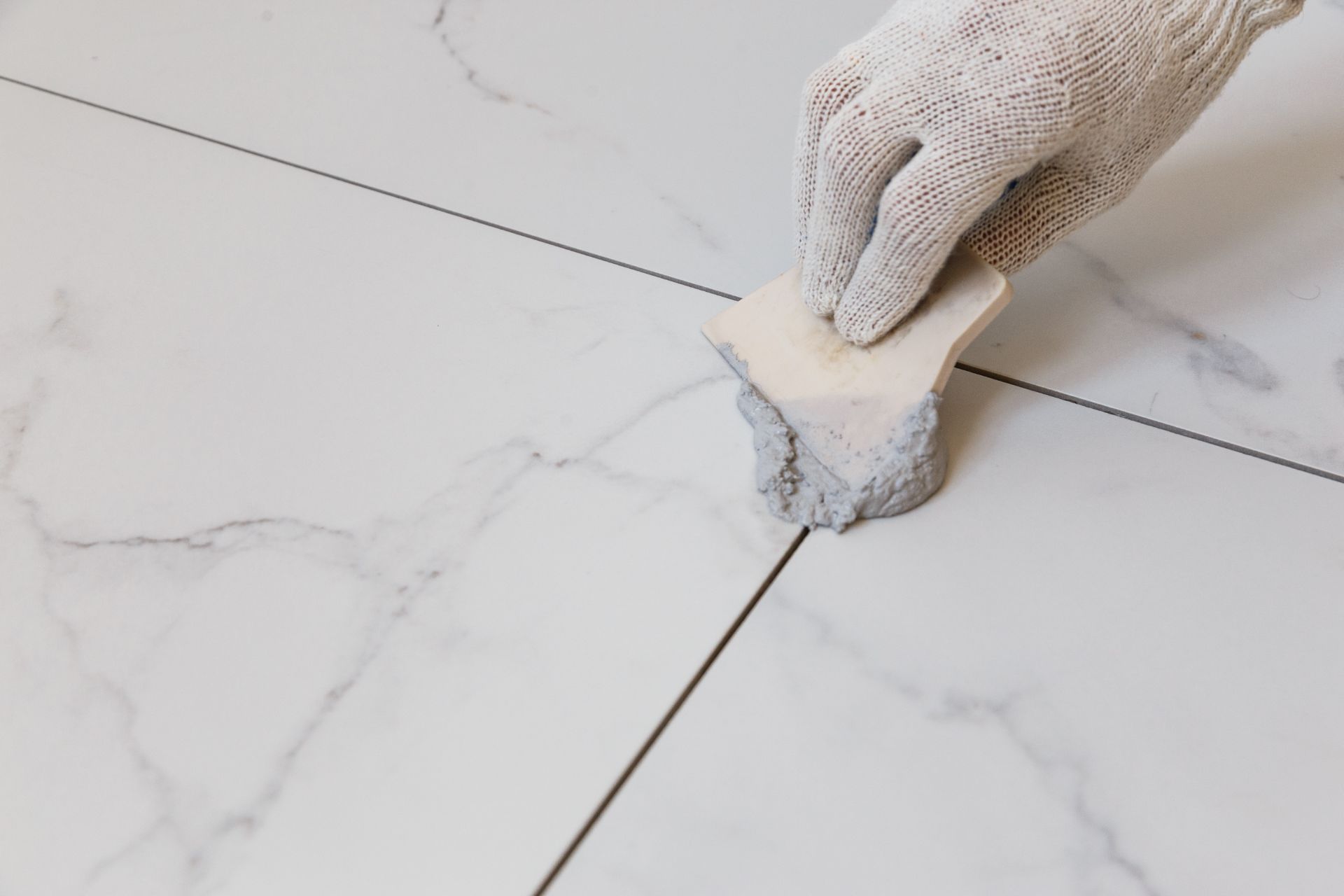Gloved hand using a grout sponge on white marble floor tiles with black grout lines.