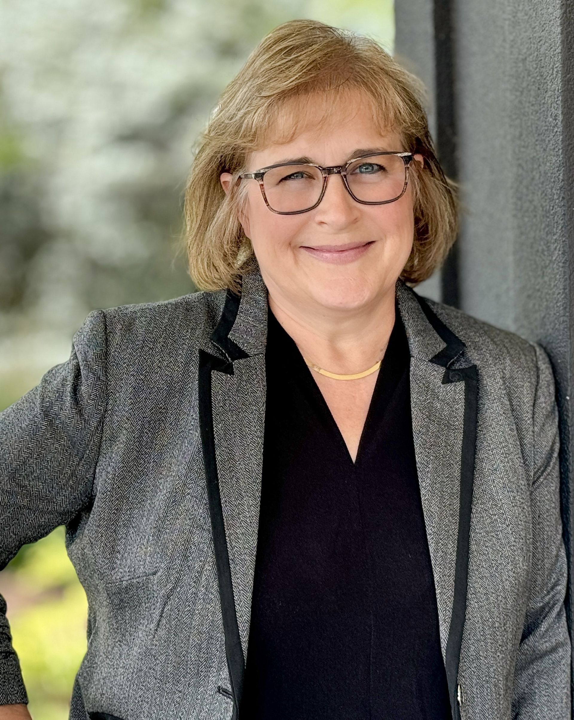 A woman wearing glasses and a gray jacket is smiling for the camera.