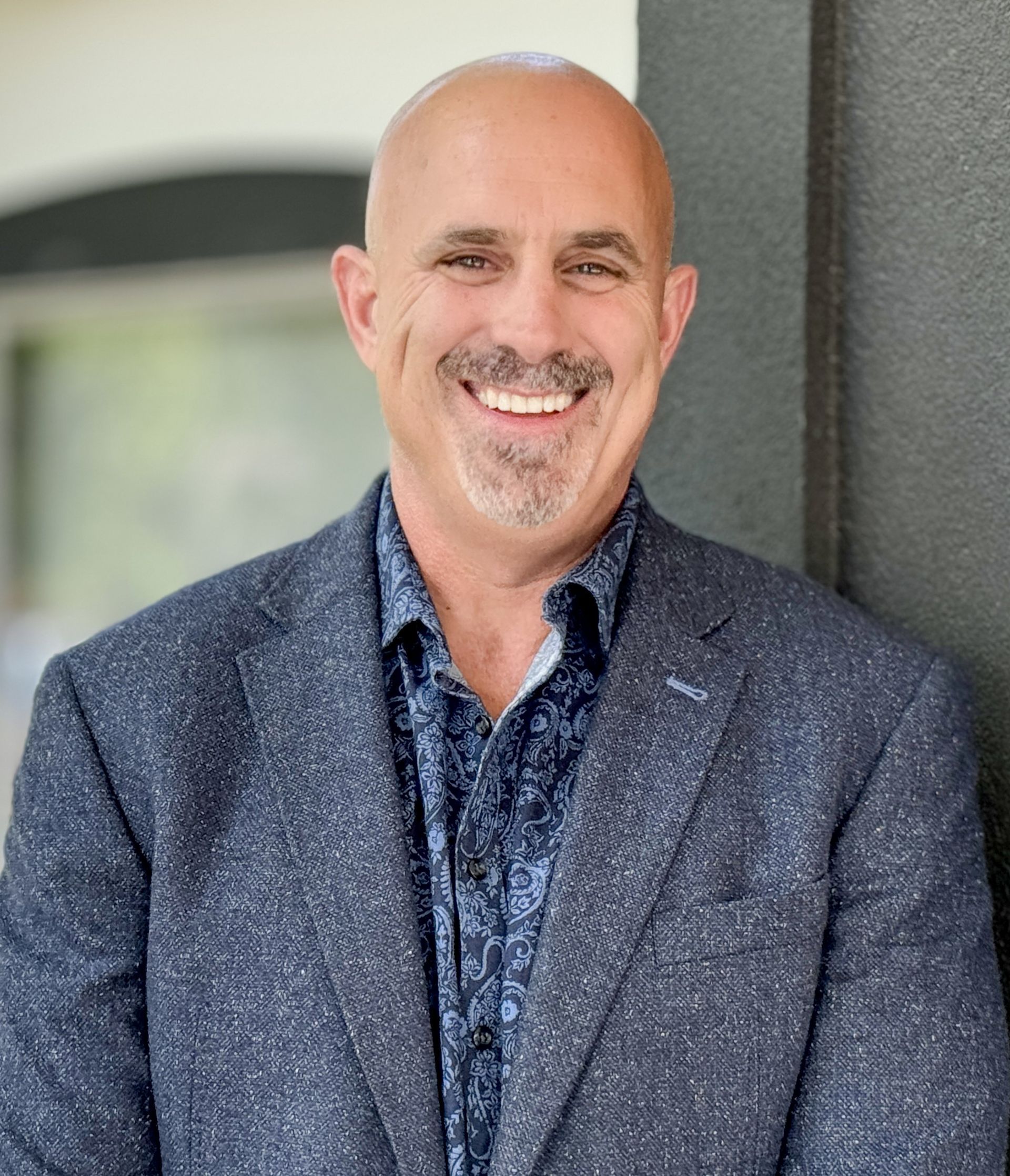 A bald man in a suit and shirt is smiling while leaning against a wall.