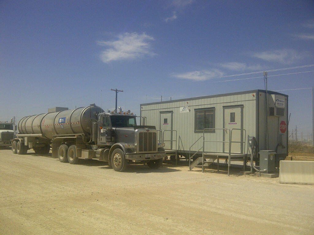 A tanker truck is parked next to a trailer in a dirt lot