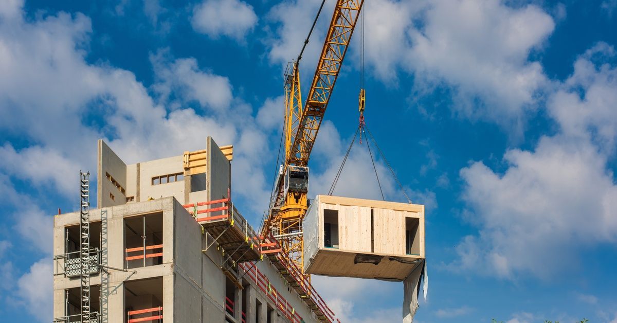 A yellow crane lifting a modular construction building section on a job site outside during the daytime.