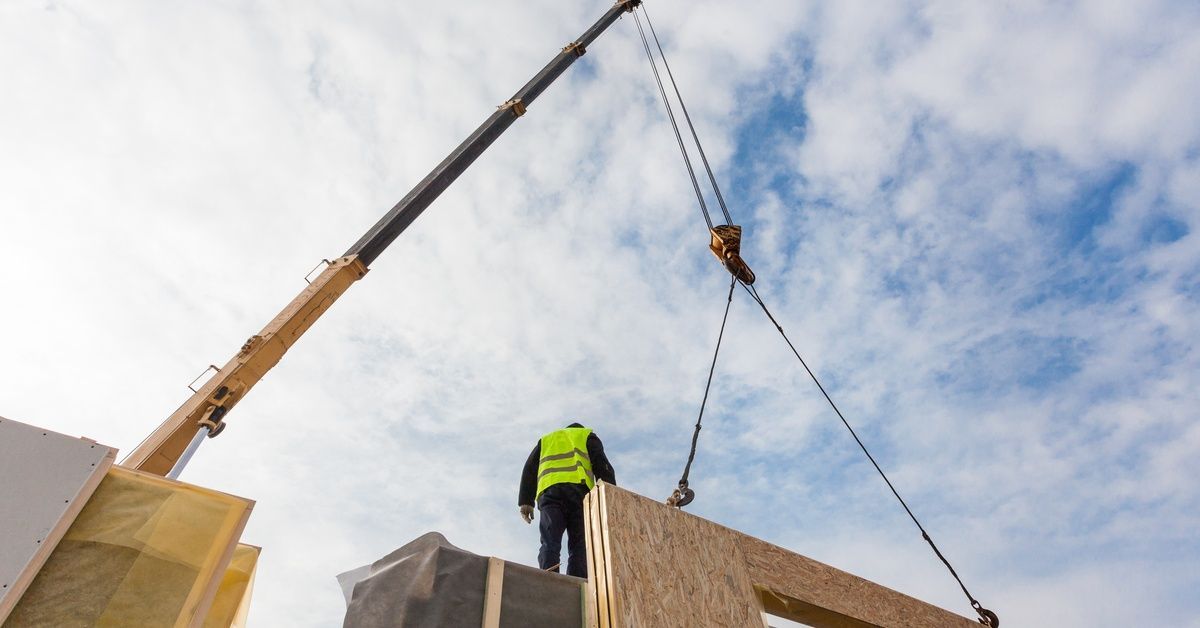A construction worker on a job site during the daytime. A crane lifts a nearby modular building section.