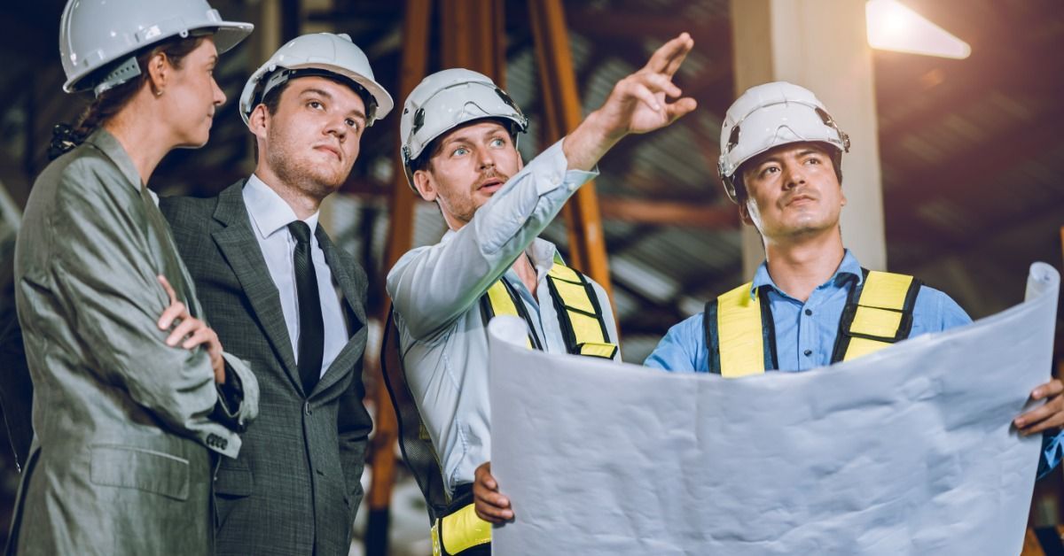 Four professionals wearing white hard hats on a construction site. One person holds blueprints.