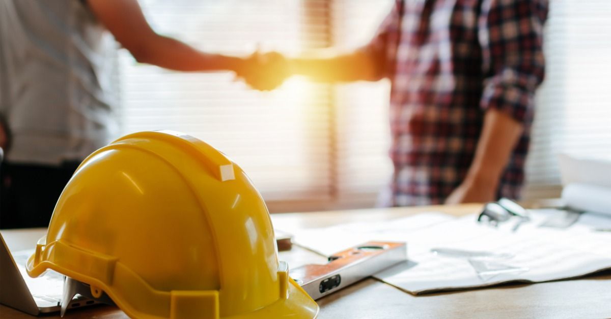 Two people shaking hands next to a window in an office. A nearby table holds a hard hat and other supplies.