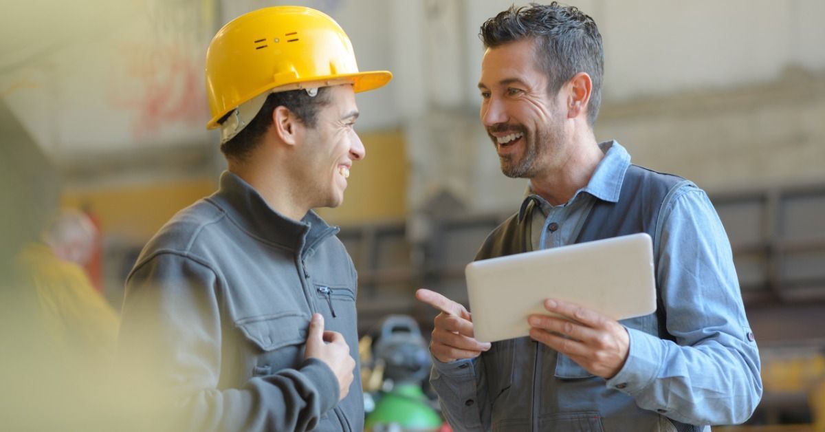 Two people laughing in a warehouse. One person wears a yellow hard hat and the other holds a tablet.