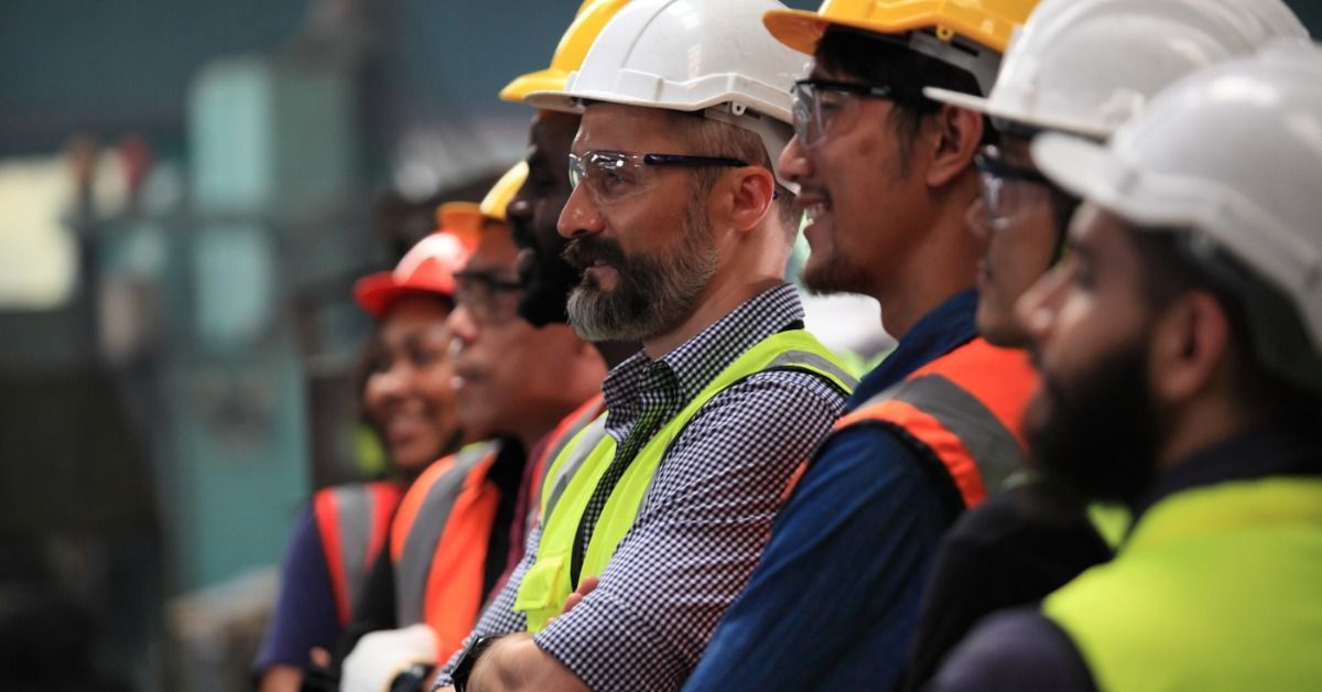 Seven construction workers standing in a large factory. Every worker wears a hard hat and a safety vest.