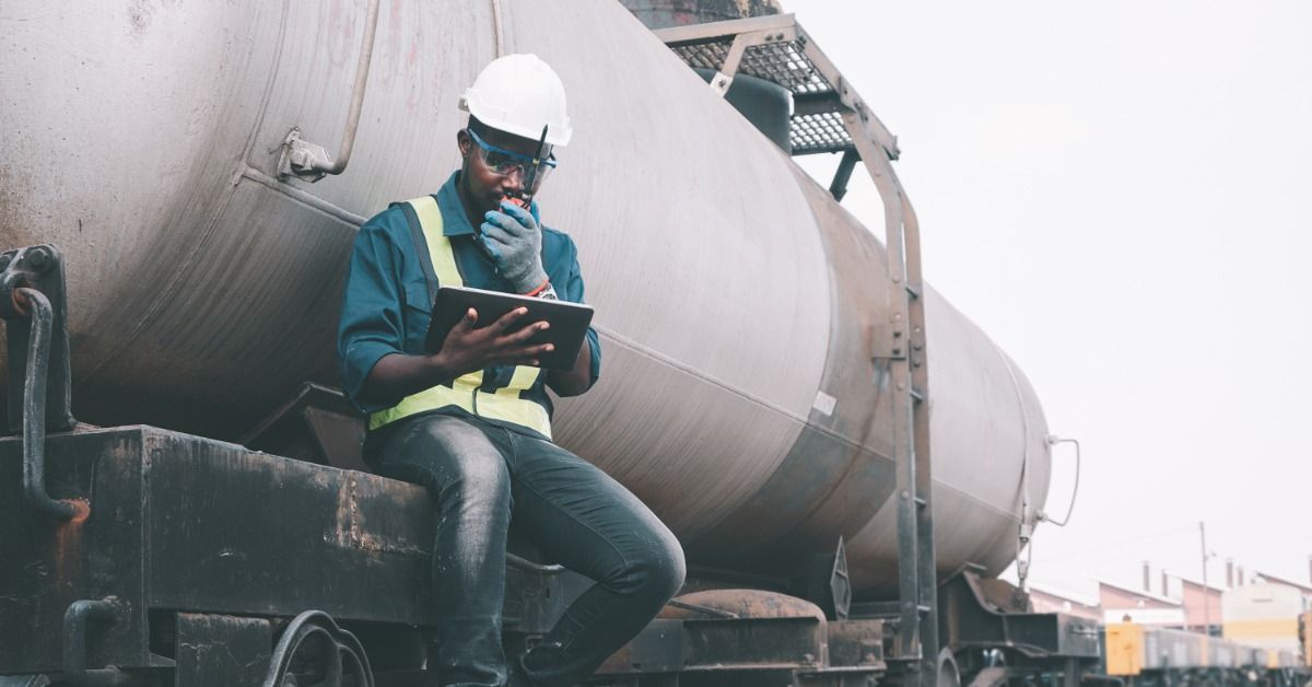 An employee sitting on an oil tank car parked on a railroad. The employee holds a walkie talkie and a tablet.