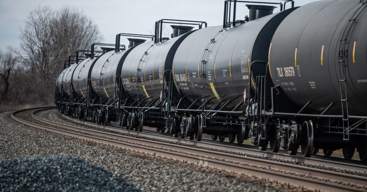 A train with seven black oil tank cars is traveling on a railroad on a cloudy day.