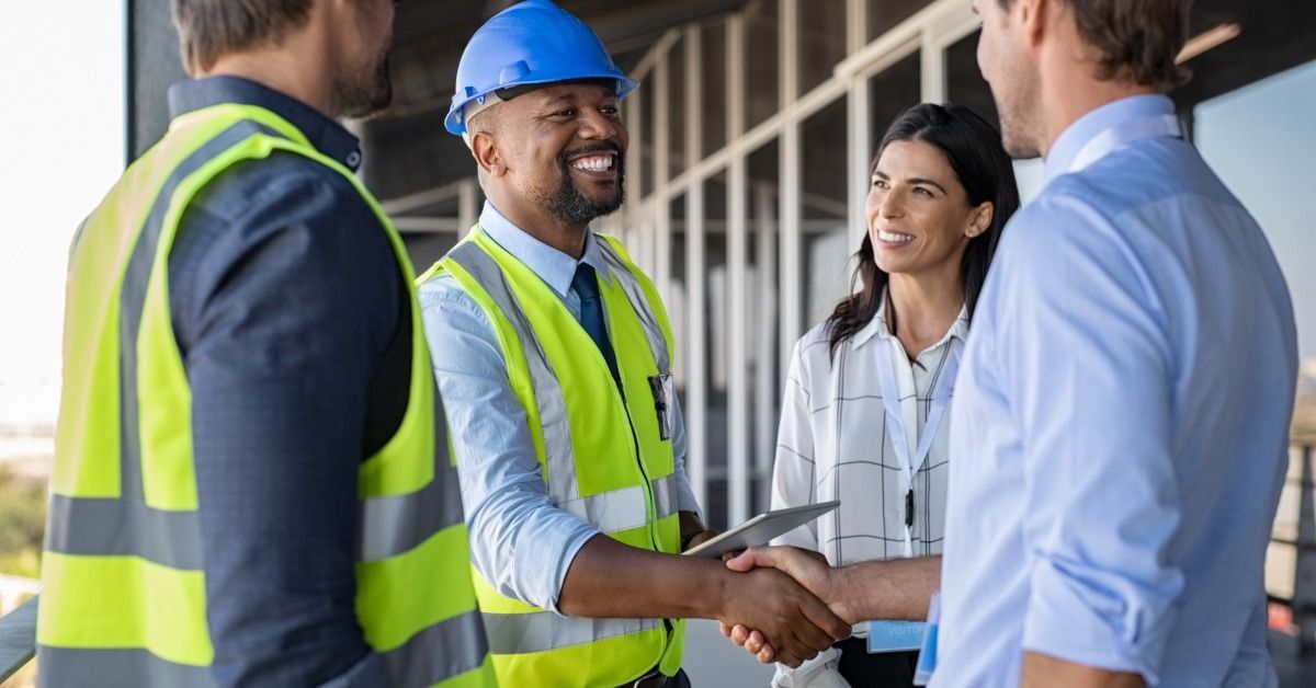 Four people standing near windows on a construction site during the daytime. Two of the people shake hands.