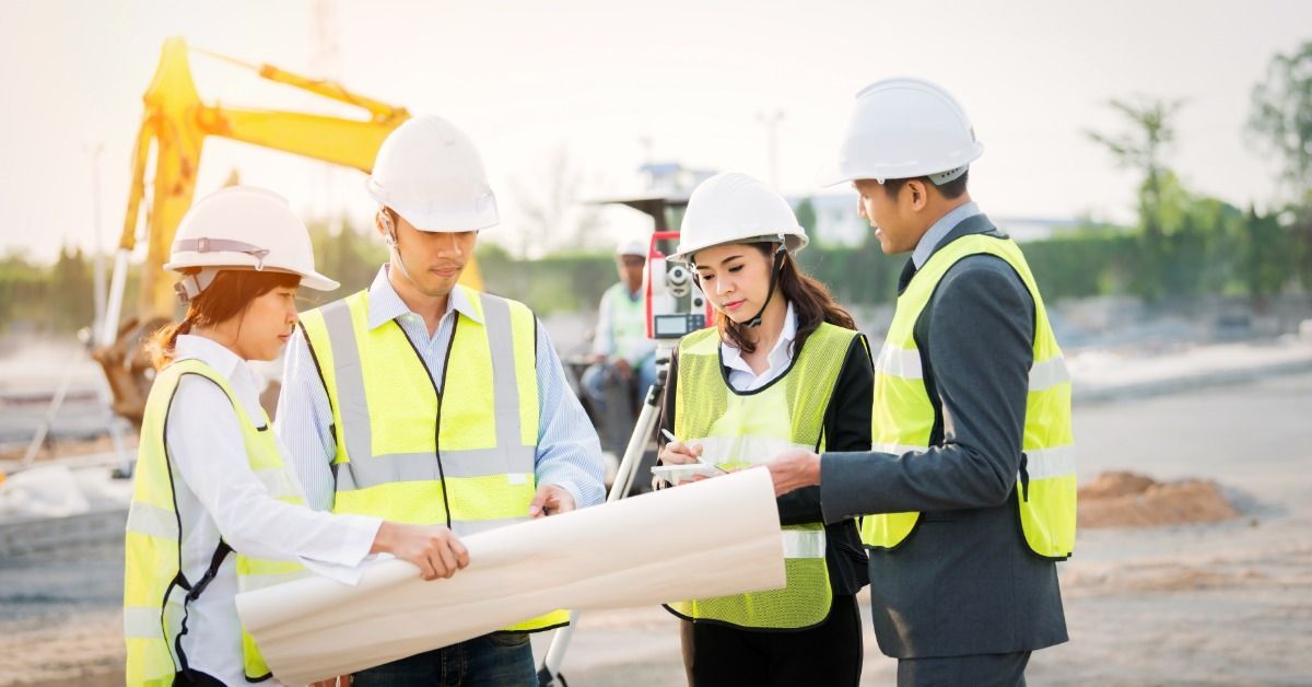 Four people wearing hard hats on a construction site. All four people look at a large set of blueprints.
