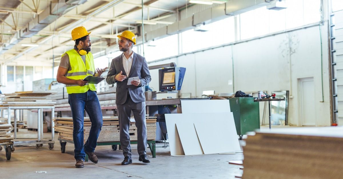 A construction worker holding a tablet and talking to a person wearing a business suit.