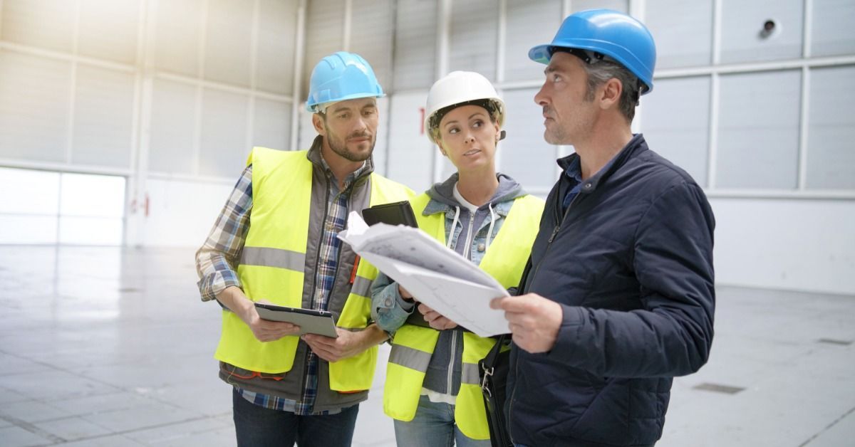 Three construction workers talking in a warehouse. Two workers wear safety vests and the third holds blueprints.