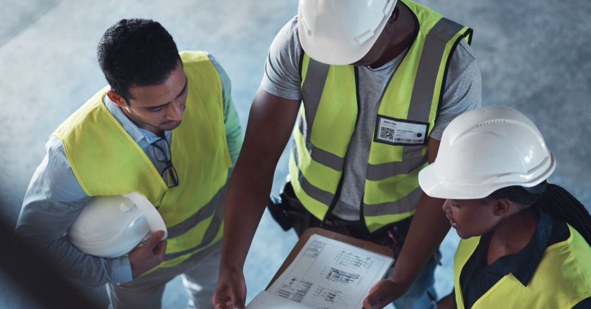 Three construction workers wearing safety vests. One worker holds a clipboard with blueprints attached to it.