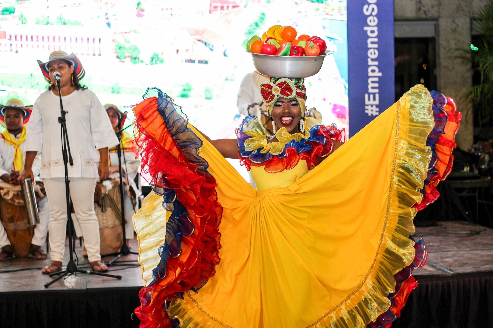 Una mujer con un vestido colorido sostiene una cesta de fruta en su cabeza. Kymbala.