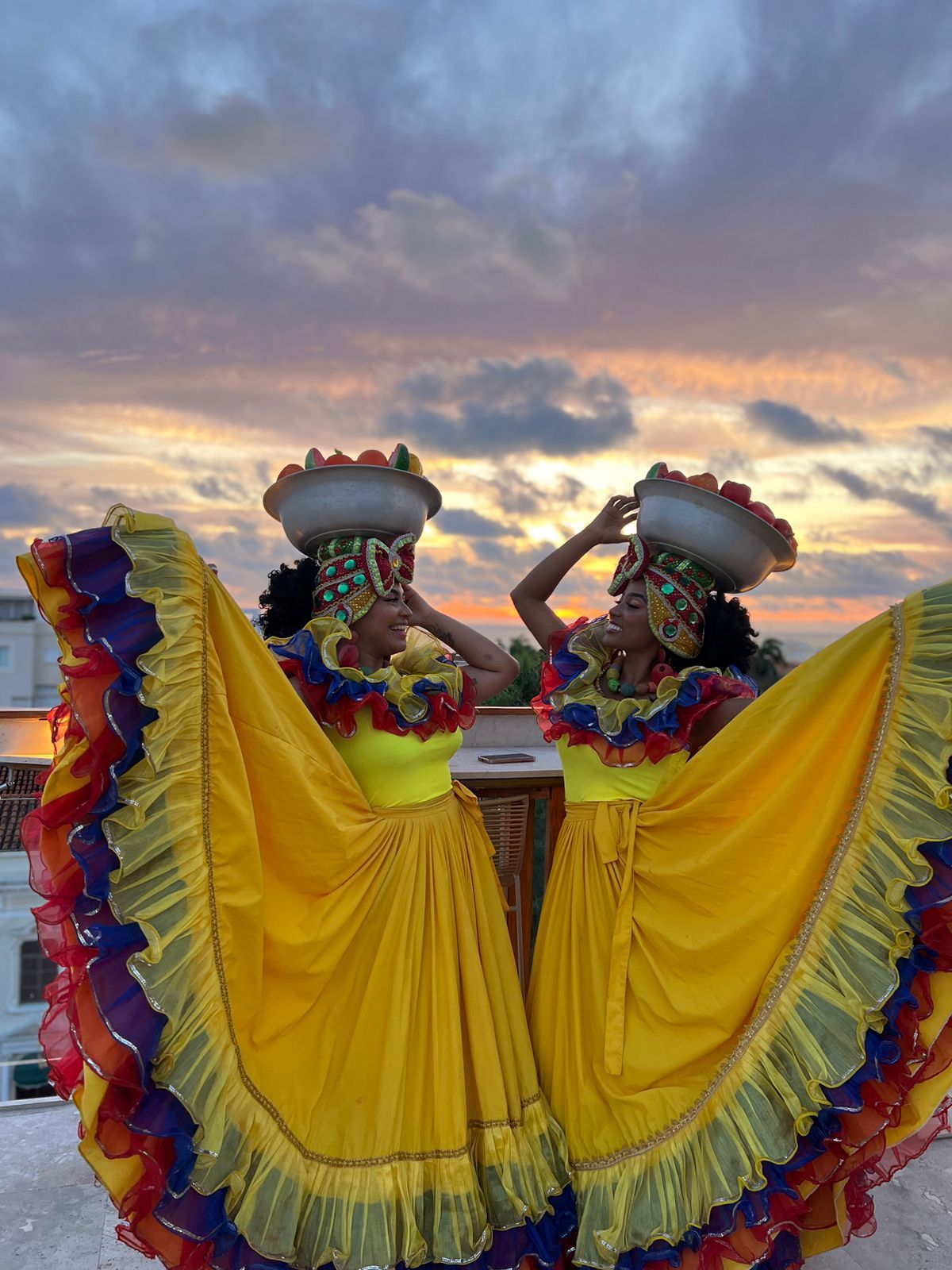 Dos mujeres con vestidos coloridos están paradas una al lado de la otra con cestas en sus cabezas.