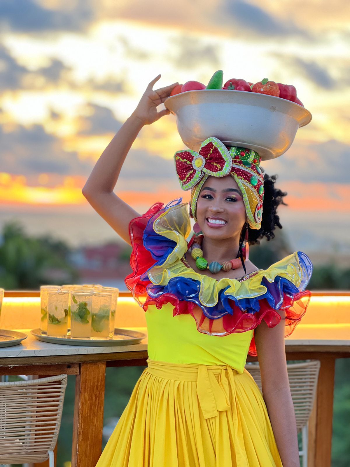 Una mujer con un colorido atuendo colombiano balancea un tazón de fruta sobre su cabeza con el atardecer como telón de fondo.